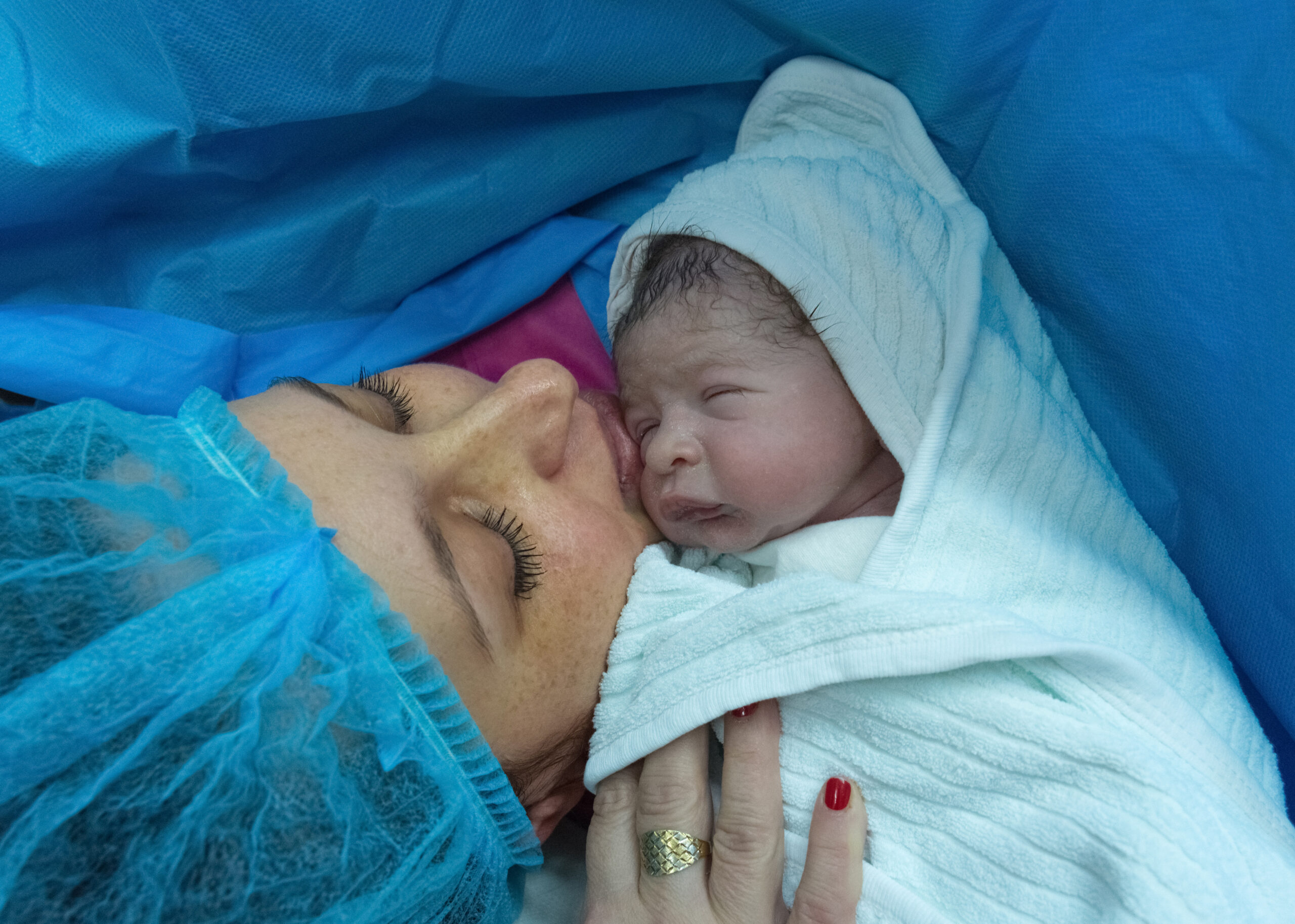 This heartwarming image captures a profoundly tender moment immediately following childbirth. A newborn baby, swaddled in a soft, light blue hooded towel, is nestled close to the mother’s face. The baby’s skin has a reddish hue typical of newborns, and dark, wet hair is visible beneath the hood. The mother, lying on her side, has her eyes closed and a gentle smile playing on her lips as she kisses the baby’s forehead. She is wearing a light blue surgical cap and gown, indicating a hospital setting, likely an operating room or recovery area after a Cesarean section. Her makeup is visible, with defined eyebrows and long eyelashes, and she’s wearing a gold ring with a large stone on her ring finger. Her fingernails are painted a vibrant red. The background is dominated by a draped, sterile blue surgical sheet, creating a calming and clinical atmosphere. A glimpse of a pink cloth can be seen near the mother’s head. The overall composition emphasizes the intimate bond between mother and child, a moment of pure joy and relief after the birthing process. The lighting is soft and natural, enhancing the emotional impact of the scene.
